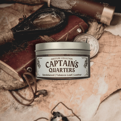 8oz Captain's Quarters candle by Emberlight Archives in a silver tin with illustrated label against a vintage table with maps background, Tobacco and Sandalwood scent.