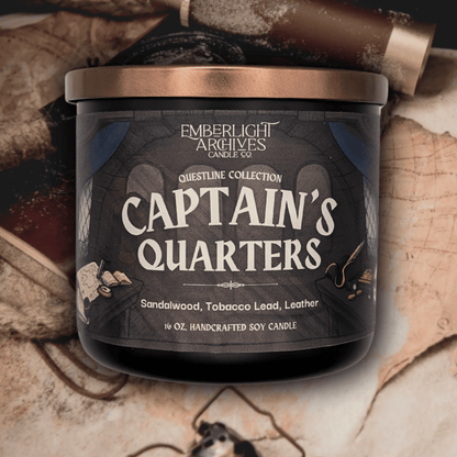 16oz Captain's Quarters candle by Emberlight Archives in a black glass jar with illustrated pirate ship label against a vintage table with maps background, Tobacco and Sandalwood scent.