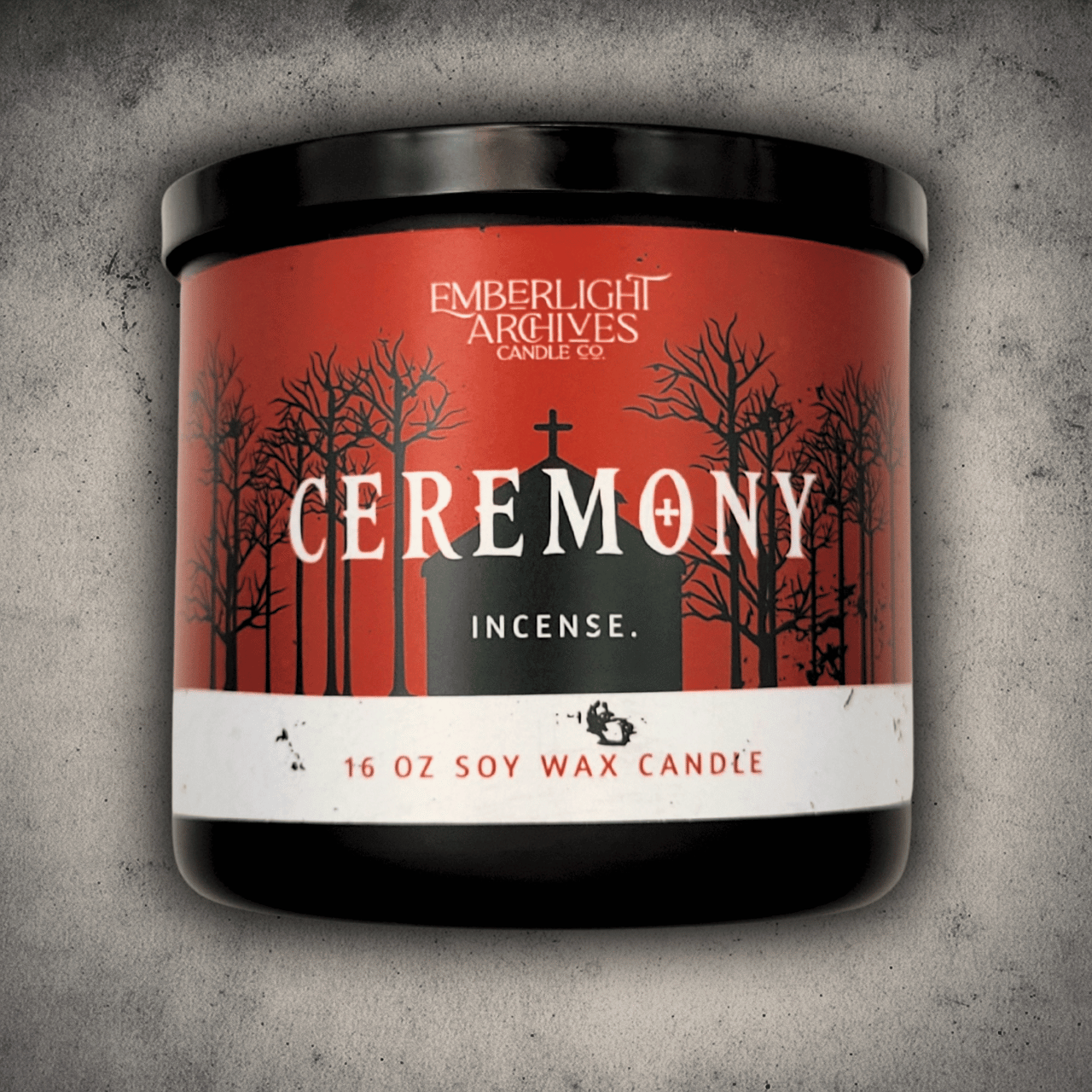 Emberlight Archives Ceremony candle in a 16oz black glass jar with red church scene label against a gray stone background, Incense scent.
