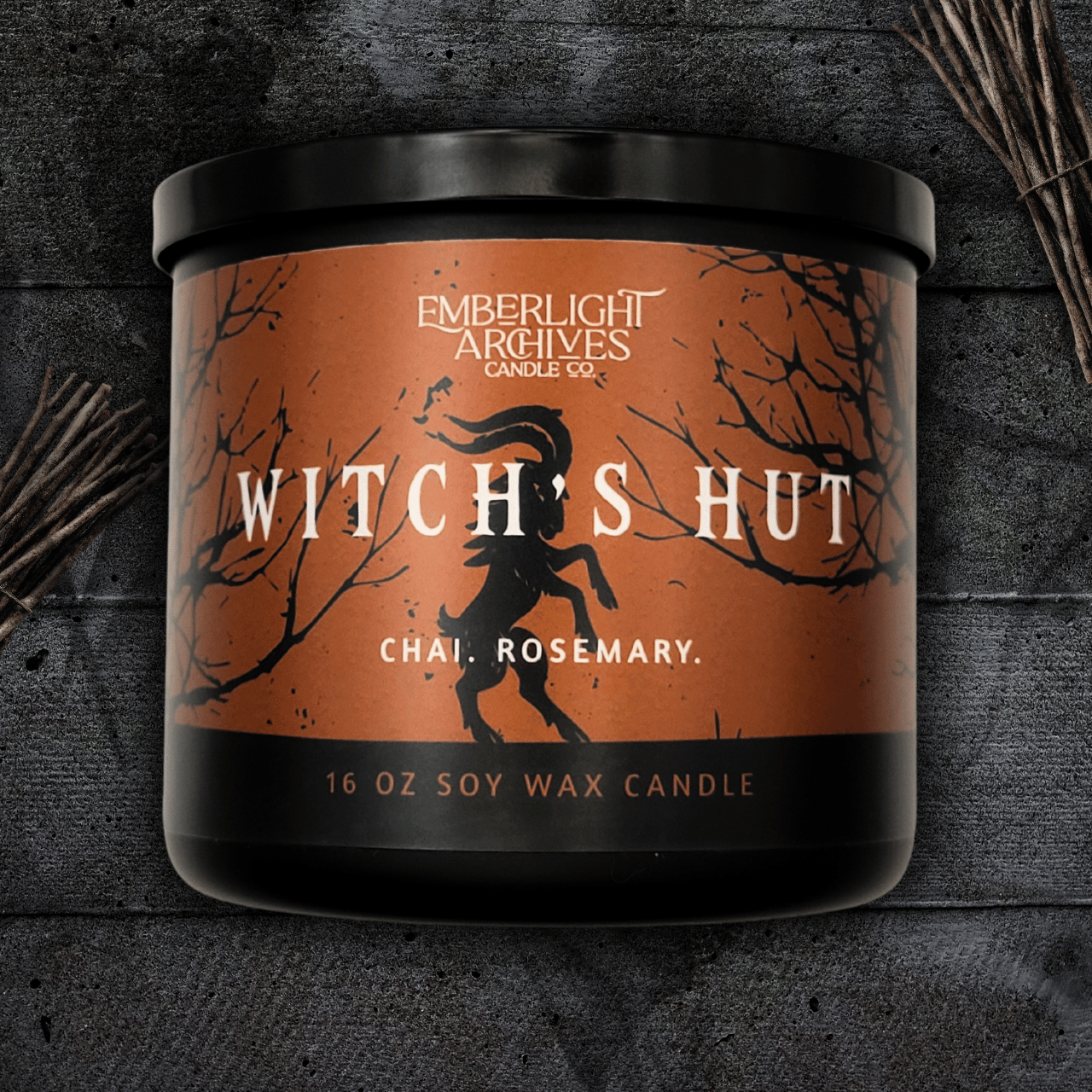 16oz Witch's Hut candle by Emberlight Archives in a black glass jar with orange dancing goat label against a dark wood background, Chai and Rosemary scent.