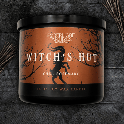 16oz Witch's Hut candle by Emberlight Archives in a black glass jar with orange dancing goat label against a dark wood background, Chai and Rosemary scent.