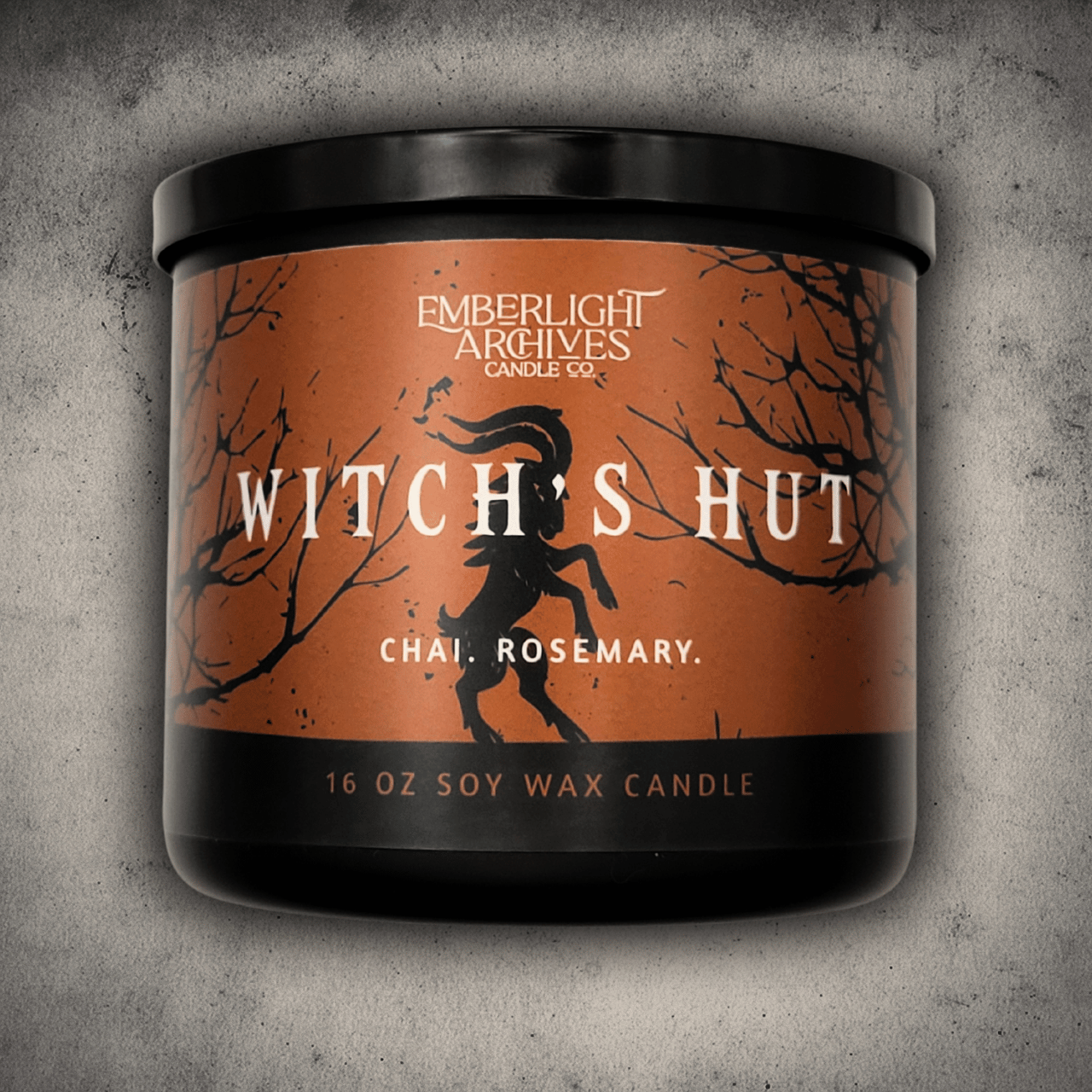 Emberlight Archives Witch's Hut candle in a 16oz black glass jar with orange dancing goat label against a gray stone background, Chai and Rosemary scent.