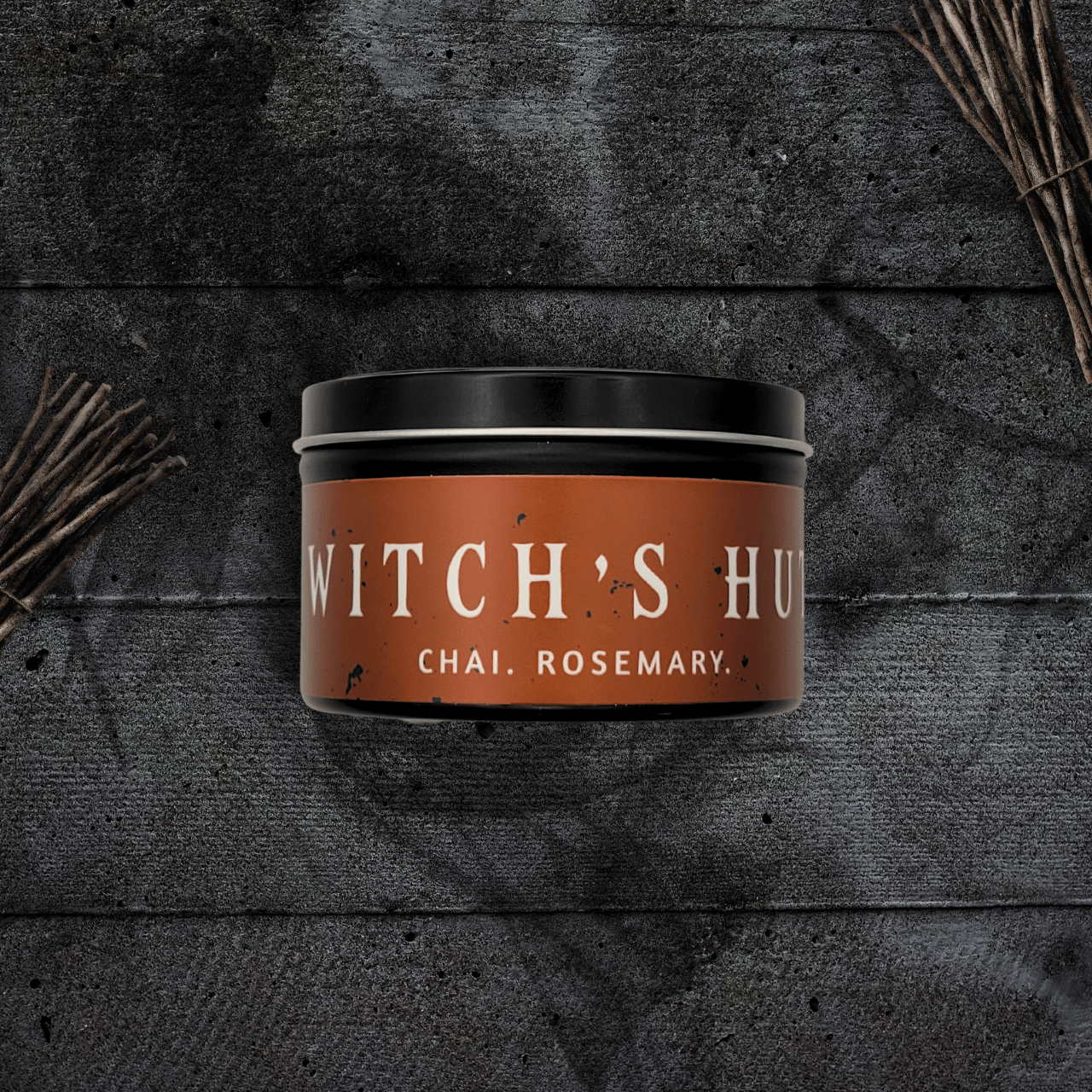 8oz Witch's Hut candle by Emberlight Archives in a black tin with orange label against a dark wood background, Chai and Rosemary scent.