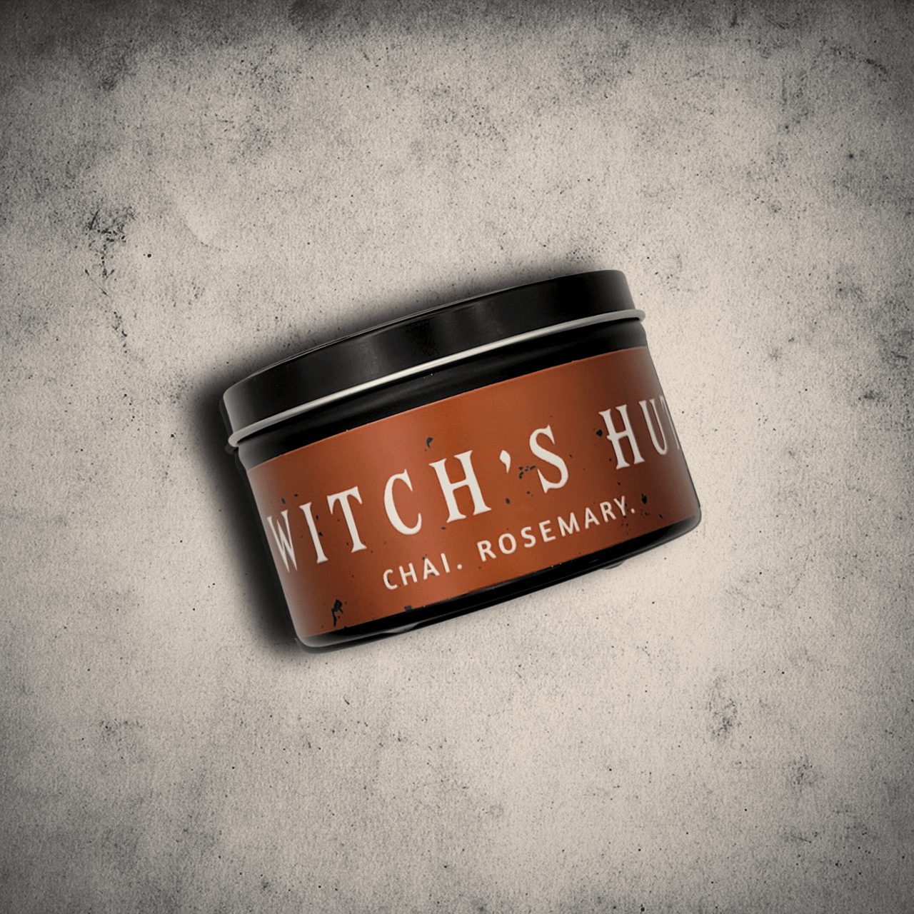 Emberlight Archives Witch's Hut candle in an 8oz black tin with orange label against a gray stone background, Chai and Rosemary scent.