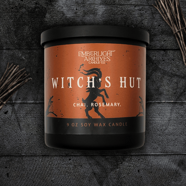 9oz Witch's Hut candle by Emberlight Archives in a black glass jar with orange dancing goat label against a dark wood background, Chai and Rosemary scent.