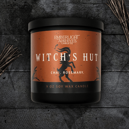 9oz Witch's Hut candle by Emberlight Archives in a black glass jar with orange dancing goat label against a dark wood background, Chai and Rosemary scent.