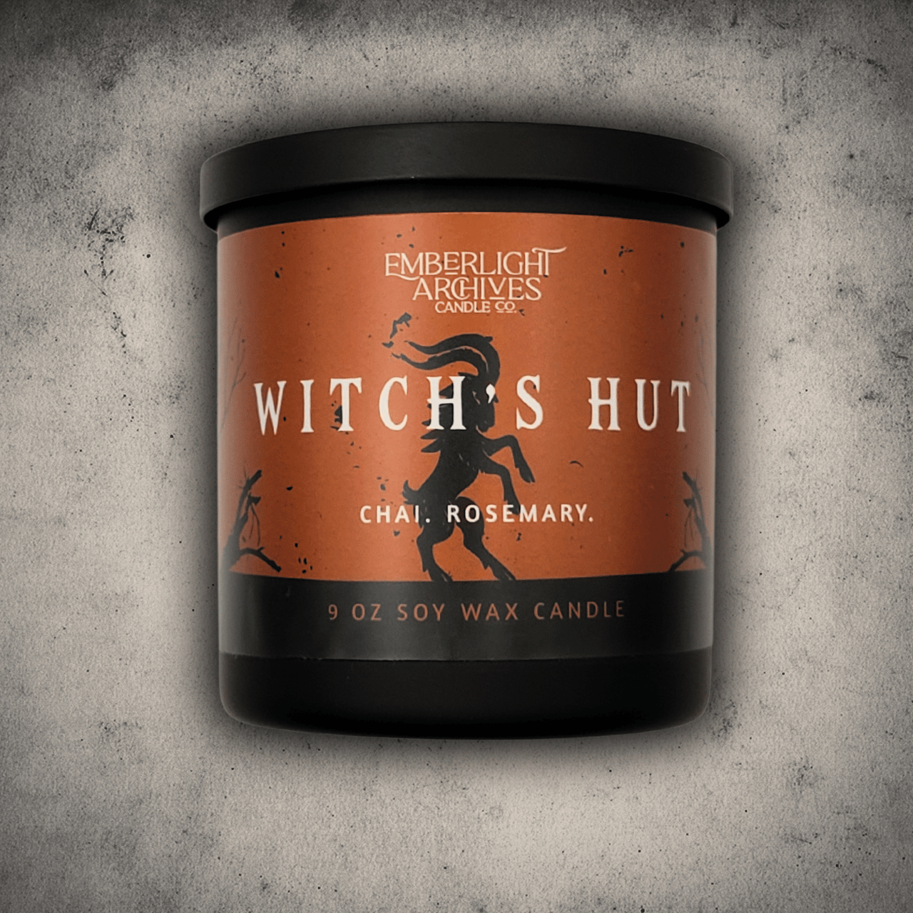 Emberlight Archives Witch's Hut candle in a 9oz black glass jar with orange dancing goat label against a gray stone background, Chai and Rosemary scent.