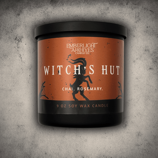 Emberlight Archives Witch's Hut candle in a 9oz black glass jar with orange dancing goat label against a gray stone background, Chai and Rosemary scent.