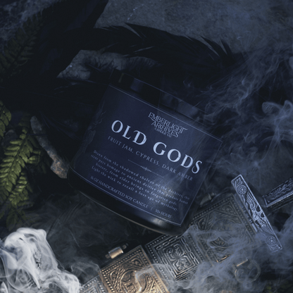 Candle labeled 'Old Gods' from Emberlight Archives on a dark background with smoke, feathers, and a sword.