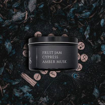 Black candle container with fragrance description reading Fruit Jam, Cypress, Amber Musk on a dark leafy background.