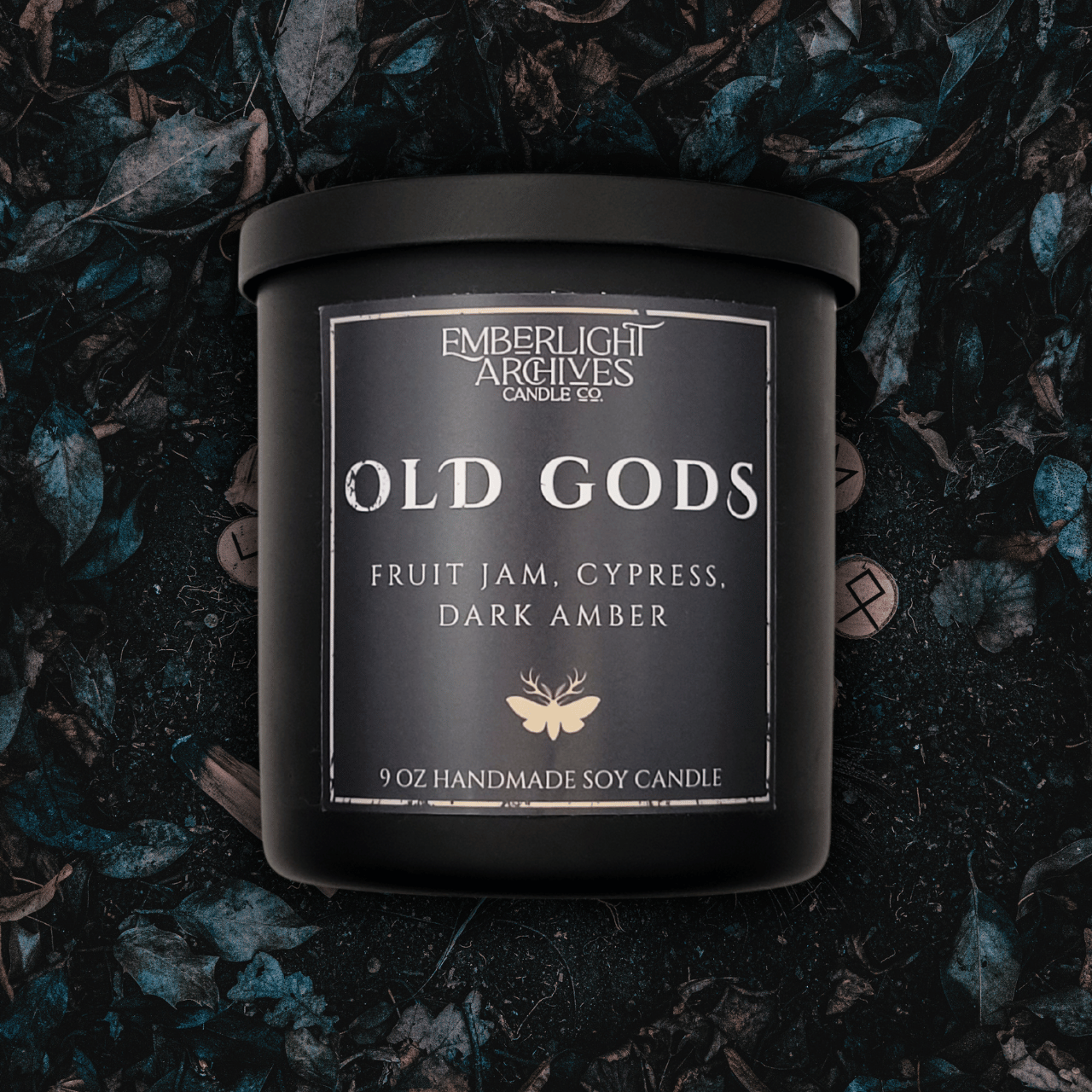 9oz Old Gods candle by Emberlight Archives in a black glass jar with dark label against a dark grassy background, Fruit Jam & Musk scent.