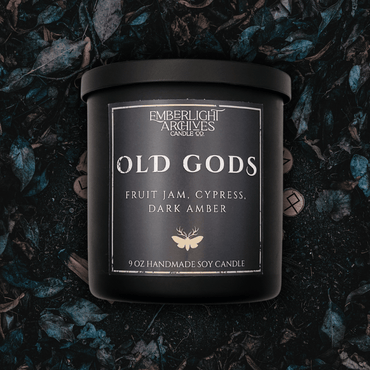 9oz Old Gods candle by Emberlight Archives in a black glass jar with dark label against a dark grassy background, Fruit Jam & Musk scent.