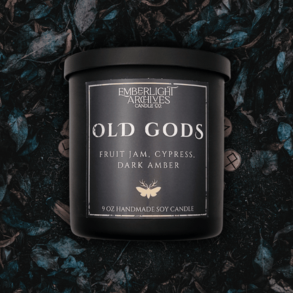 9oz Old Gods candle by Emberlight Archives in a black glass jar with dark label against a dark grassy background, Fruit Jam & Musk scent.